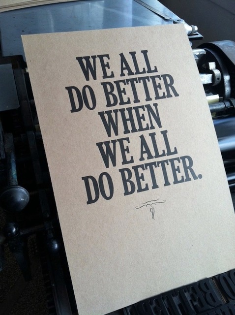 A brown rectangular poster is propped up on a letterpress machine. It reads We All Do Better When We All Do Better in black serif typeface. 