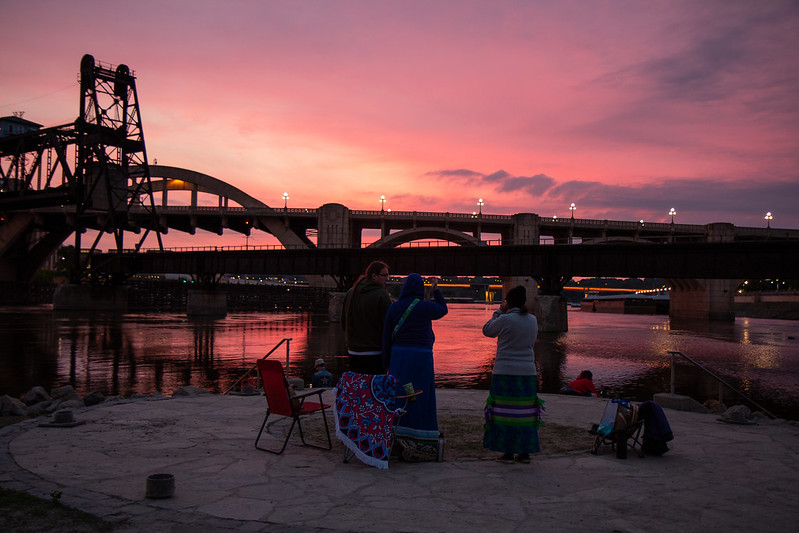 A vibrant pink and purple sunrise behind bridges and the river. In the foreground, people stand and admire the sky.