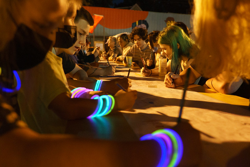 A crowd of people sit along a table outside at night. All of them hold pencils in their hands and focus on writing and drawing. Some wear glowing bracelets and necklaces.