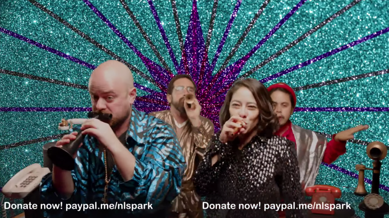 Four people in sparkly outfits play kazoos of all shapes and sizes in front of a green screen displaying a turquoise, purple, and black sequin explosion design radiating out from the center.