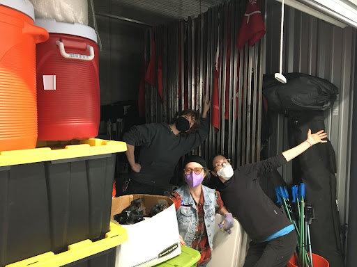 Three white people with face masks pose amidst piles of bins, water cooler jugs, boxes, stakes, and other equipment.