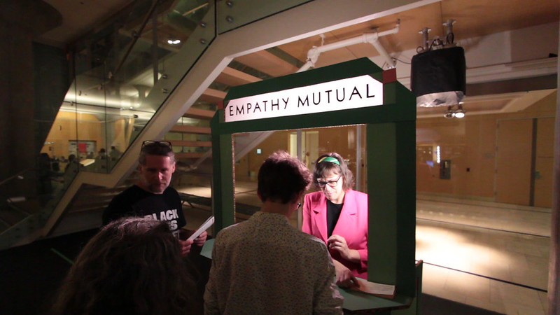 A florescent-lit green bank teller booth stands beneath the stairs of a library. The top of the booth reads, "Empathy Mutual." A person in a bright pink blazer stands in the window of the booth, and three people stand around the booth.