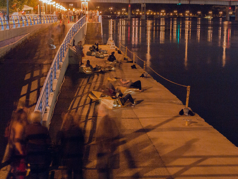 People lounge on wooden structures on a concrete landing alongside the water of the river. A coppery glow rests on the scene, and golden, red, and blue-green lights reflect on the water.