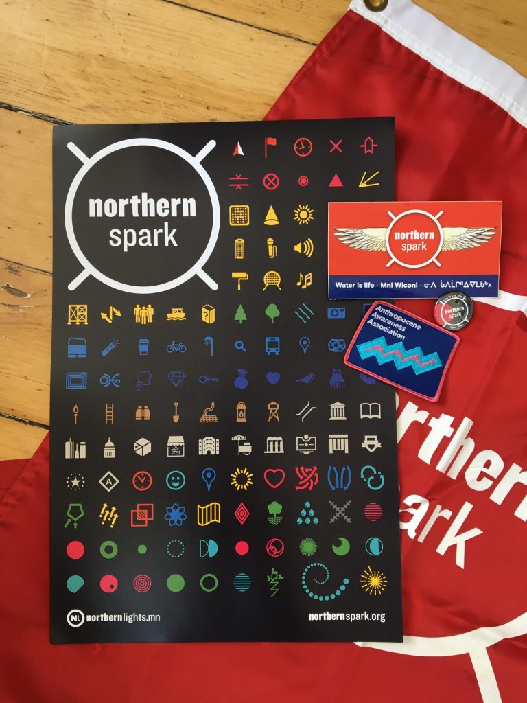An overlapping assortment of items, including a poster with colorful icons on a black background, a blue patch, a red sticker, a black and red button, all on top of a red Northern Spark flag.
