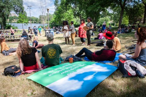 On a bright summer day in a park wtih green leafed trees, people sit on the ground on a blanket printed with a poem in white text on green and blue background. The people's attention is tuned to four people standing and reading poetry outloud. 