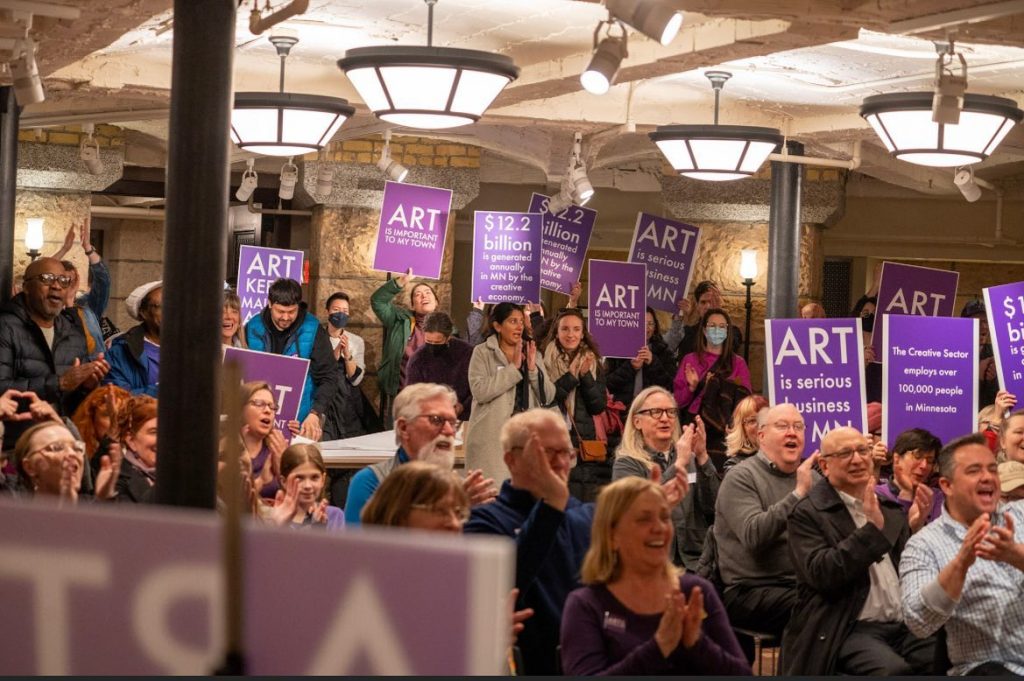People fill seats and stand around the perimeter of a room with stone and brick pillars, cheering and holding purple signs that read "Art is important to my town," and "Art is serious business in MN," among other messages.