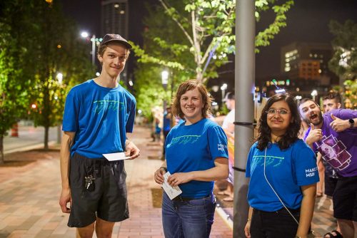 Three people wearing blue Northern Spark t-shirts stand on a sidewalk at night and smile at the camera.