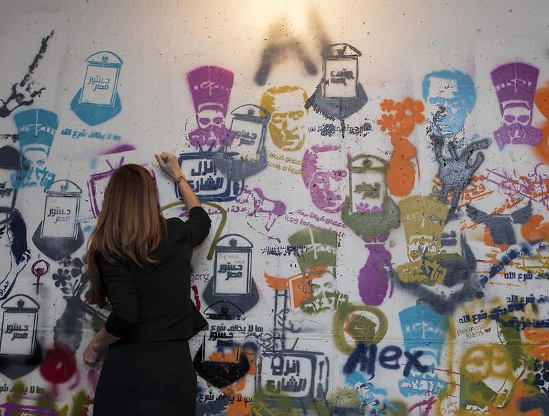 A person with their back to the camera writes on a wall covered in colorful spray-paint stenciled images from the Arab Spring, including Arabic script, faces of prominent political figures, faces behind gas masks, and television sets.