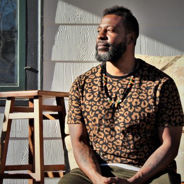 Bayou, a black man with a beard, wearing a brown and black printed shirt and black necklace, sits in the sun and smiles thoughtfully.