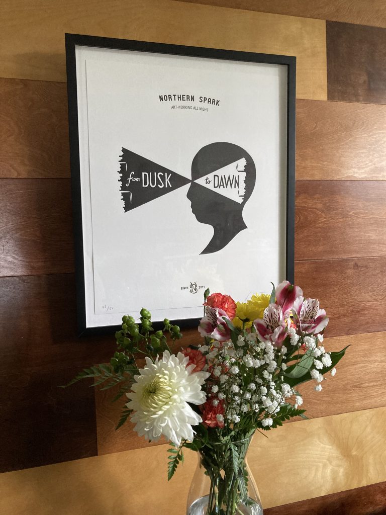 A framed image of a black-and-white letterpress print of a head in profile, with projections of a city along the edges and the words "from dusk to dawn" hangs on a wall behind an arrangement of flowers.