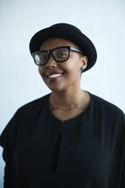 Matshoshi, a black woman wearing a matching outfit of a black brimmed hat, black glasses, black circle earrings, and black shirt, smiles at the camera.