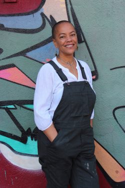 Sayge, a black woman wearing a white shirt and dark green overalls, stands with her hands in her pockets and smiles.
