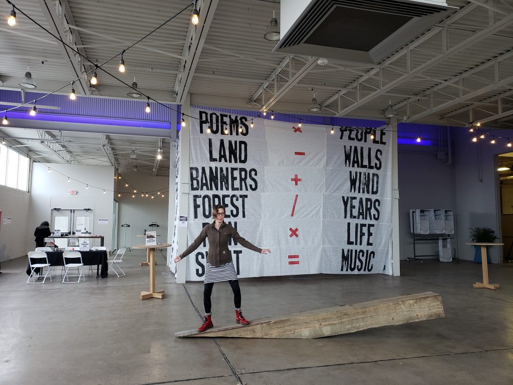 NS Producer Erin Lavelle balances on Dream the Combine's Barely Holding It Together in front of Piotr Syhalski's Living Banners. Spring Howl 2019 